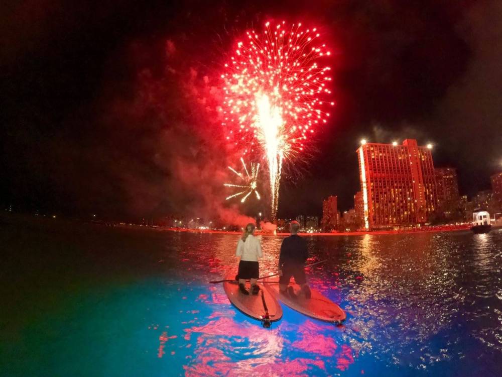 Two people on paddleboards at night under red fireworks near city buildings.