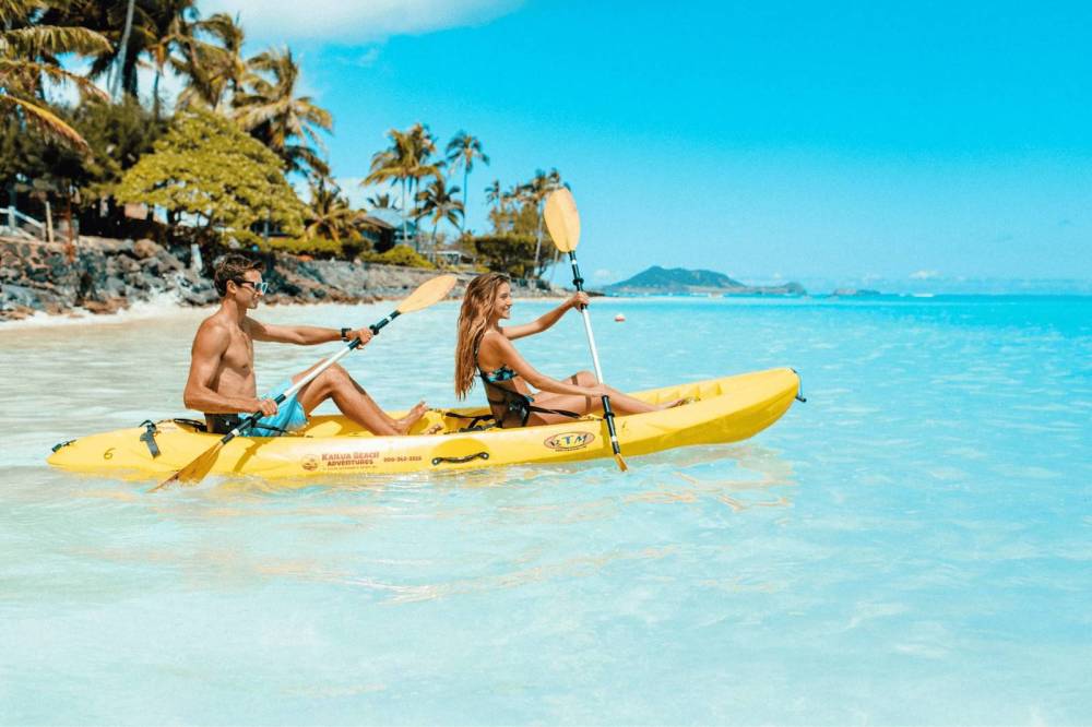 Two people kayaking in a yellow kayak on clear blue water with palm trees in the background.