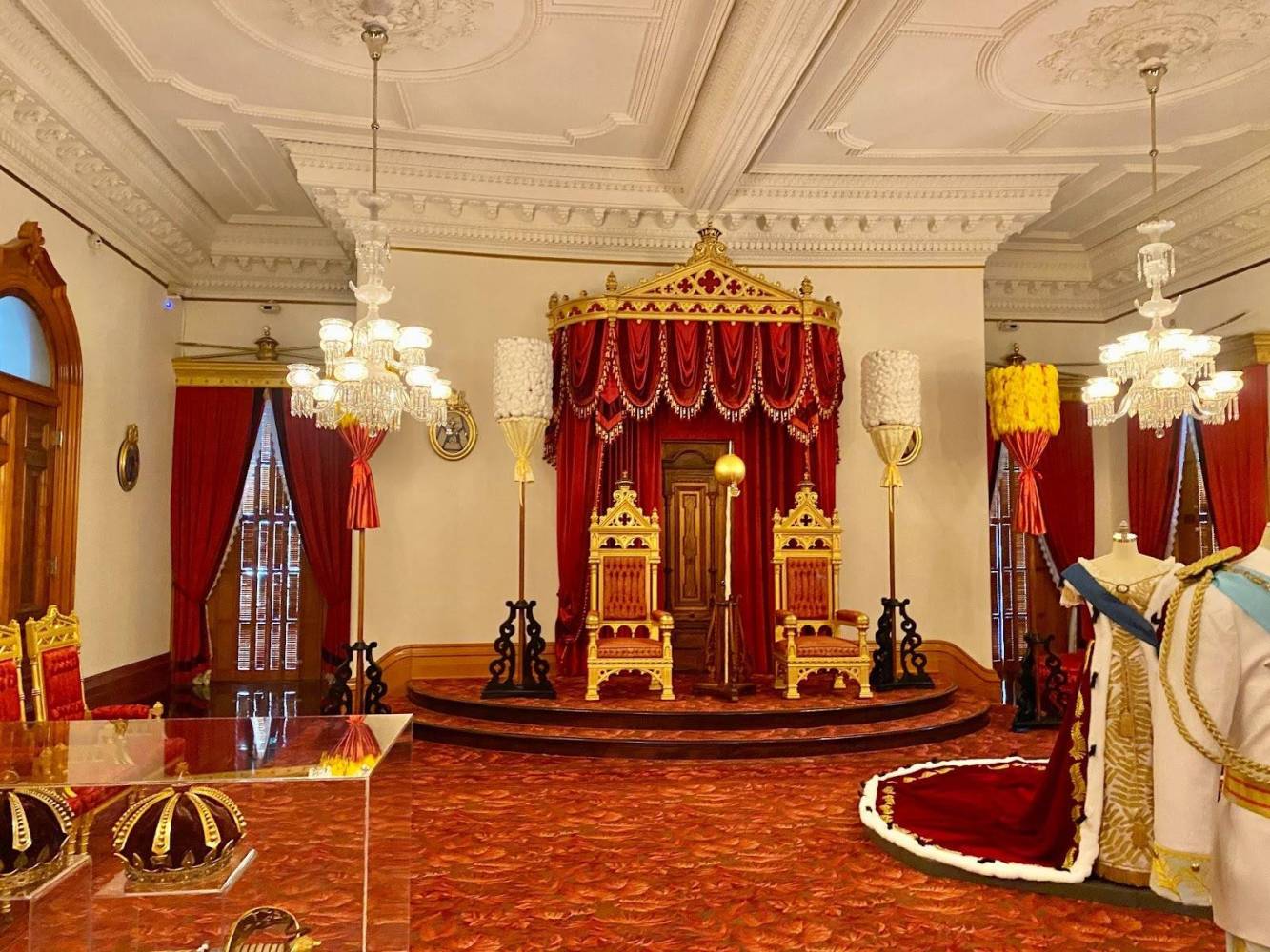 Opulent throne room with red and gold decor, featuring two ornate thrones and chandeliers.