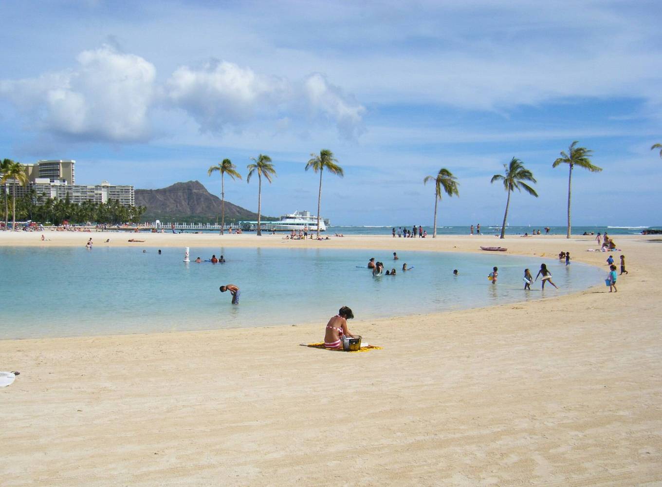 Beach scene with people, palm trees, and distant mountain under a blue sky.