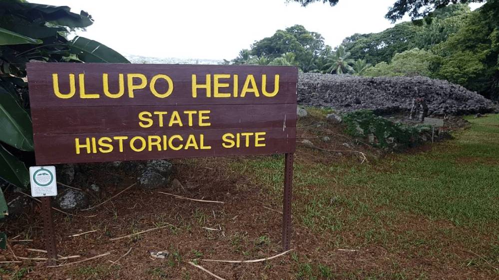 Sign reading 'Ulupo Heiau State Historical Site' with stone structure and trees in the background.