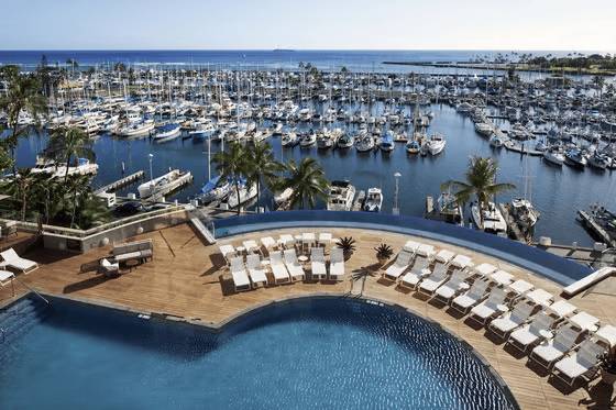 Aerial view of a marina with boats, a circular pool, and lounge chairs on a deck overlooking the ocean.