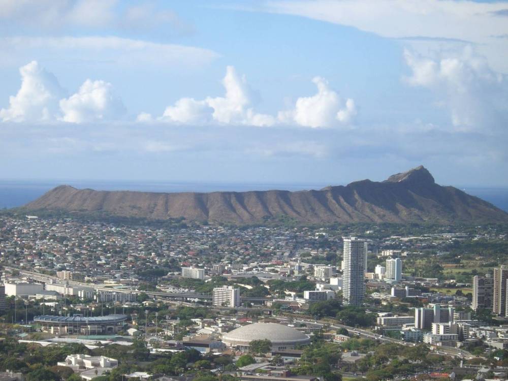 Aerial view of a cityscape with a distinct mountain in the background under a partly cloudy sky.