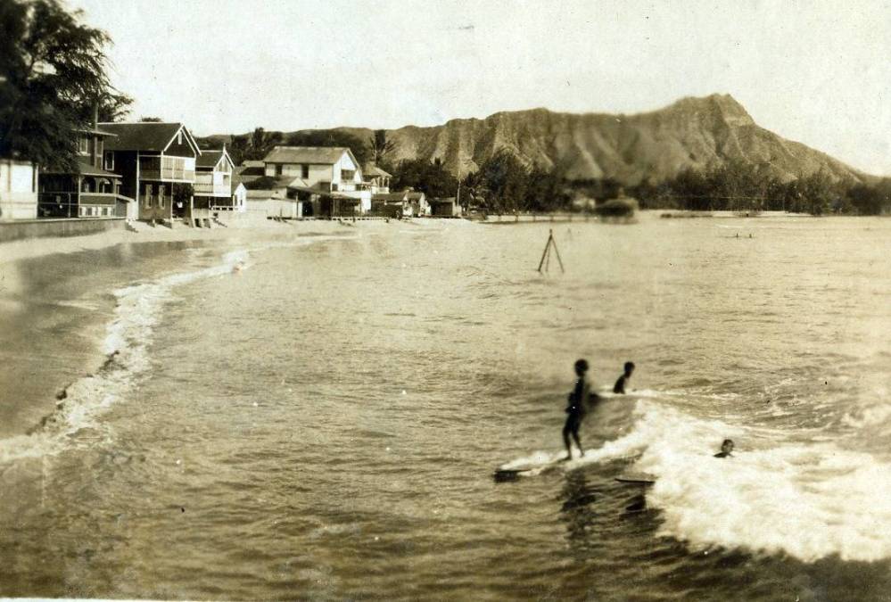 Vintage photo of surfers near a beach with houses and a mountain in the background.