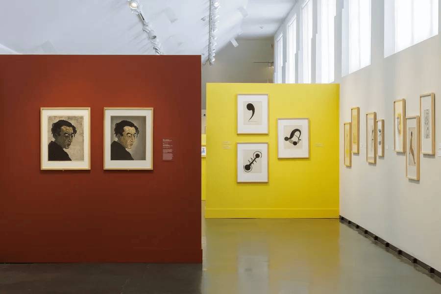 Art gallery with framed artworks on red and yellow walls, featuring abstract and portrait pieces.