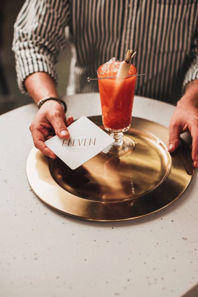 Person placing a drink and a card with 'ELEVEN' on a gold platter.