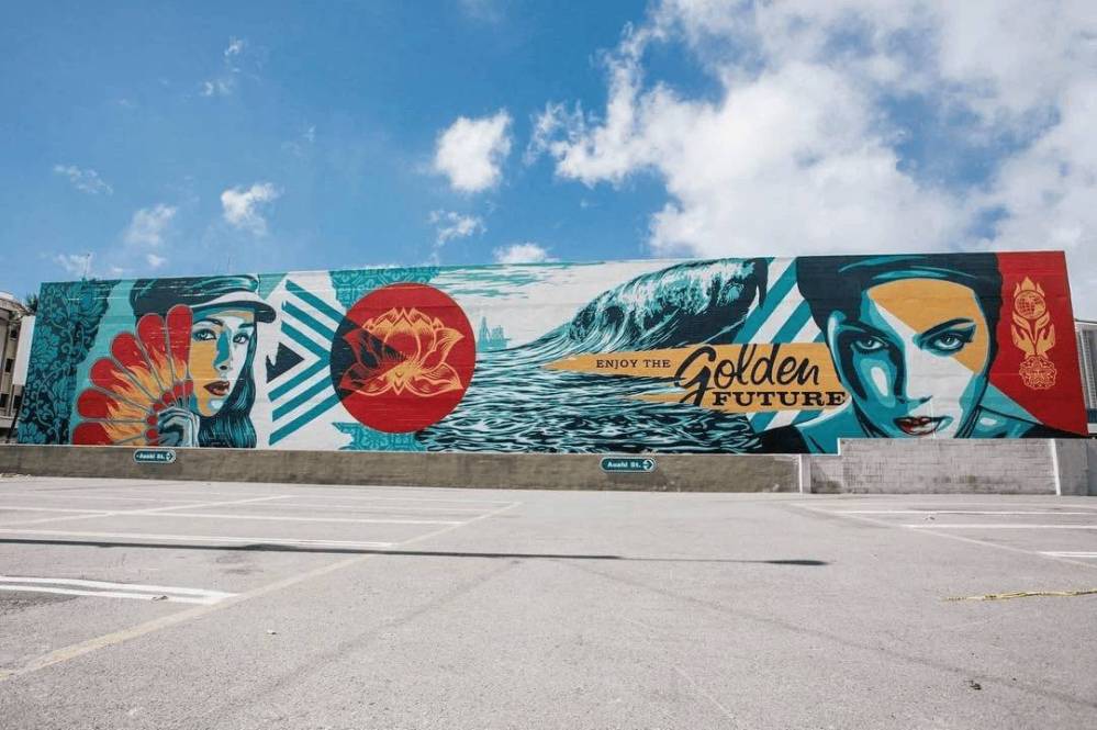 Colorful mural with faces, waves, and 'Enjoy the Golden Future' text on a building under blue sky.