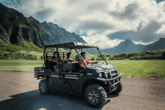 People in an ATV driving on a dirt path through scenic mountains and grass under a cloudy sky.
