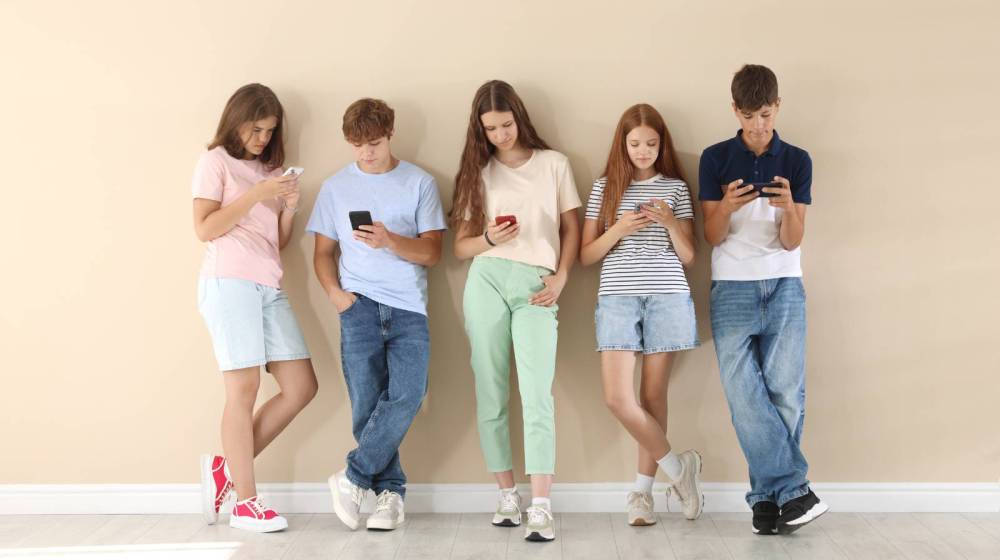 Five teenagers standing in a row, looking at their smartphones against a beige wall.