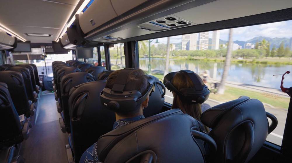 Bus interior with passengers wearing VR headsets, window view shows lake and cityscape.