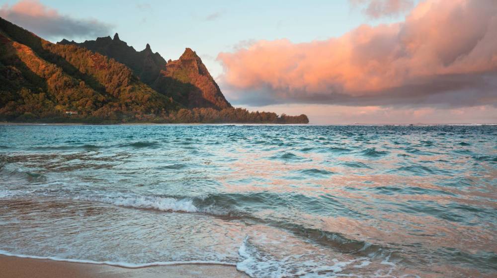Coastal scene with waves, beach, mountains, and pink clouds at sunset.