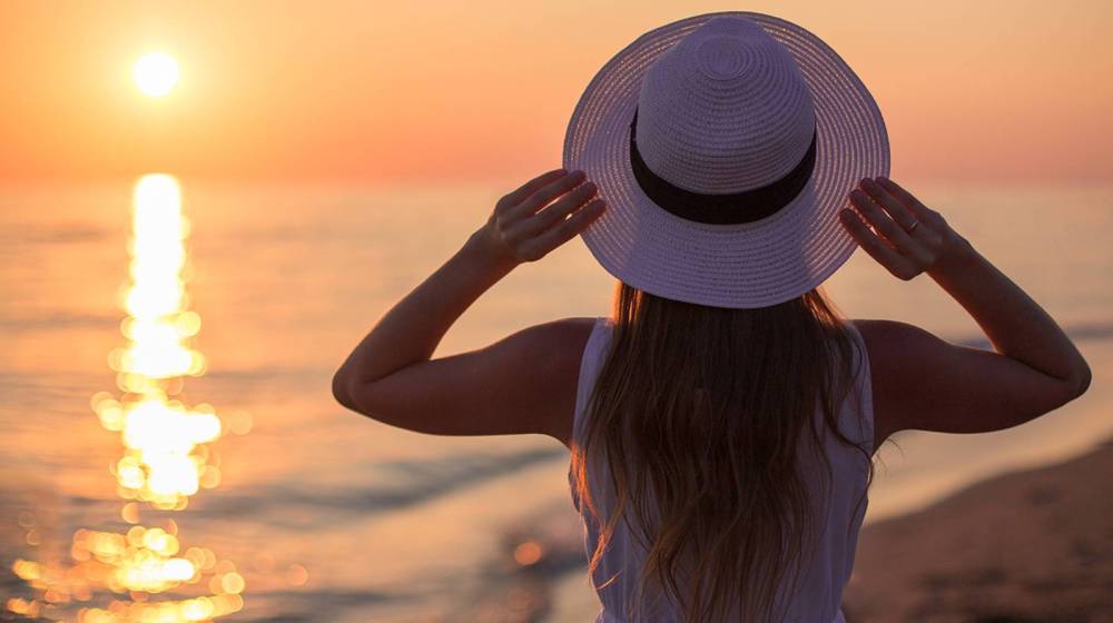 Person with hat faces ocean sunset, holding brim with both hands on a sandy beach.