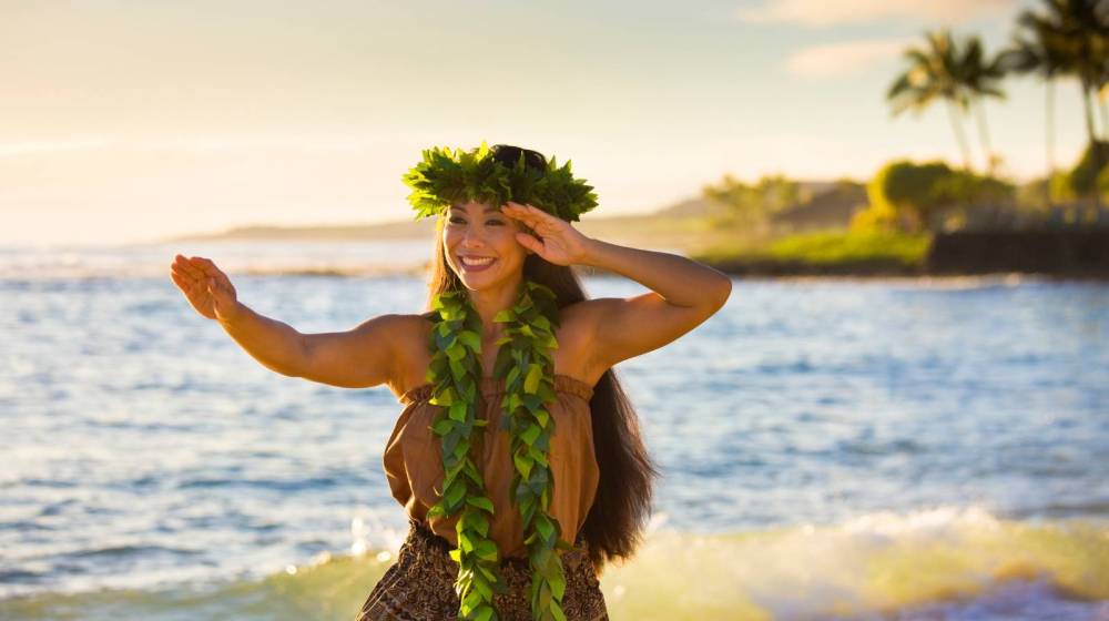 A woman in traditional attire dances on a beach with a leafy headpiece.