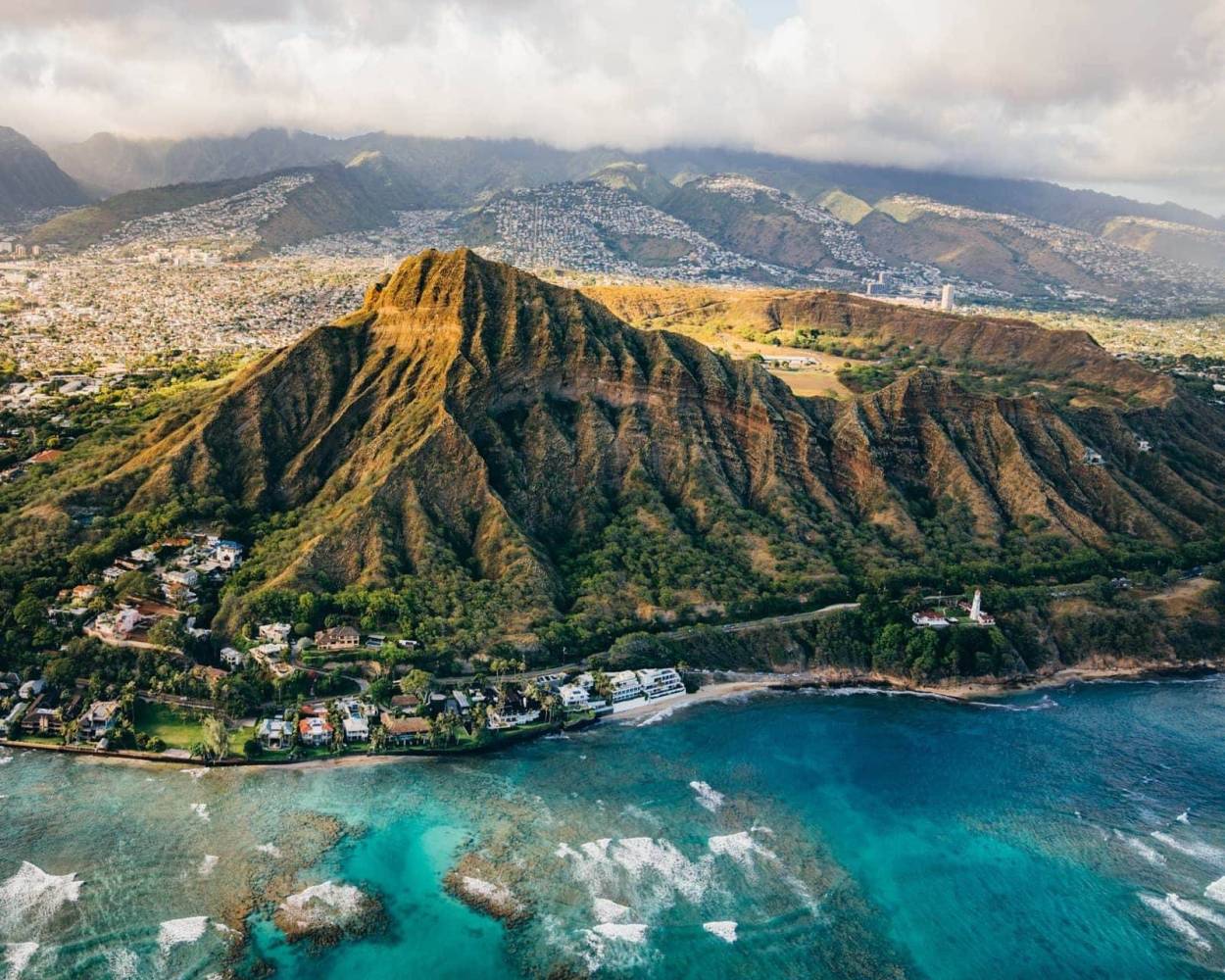 Aerial view of Diamond Head crater with blue ocean in foreground and cityscape in background.