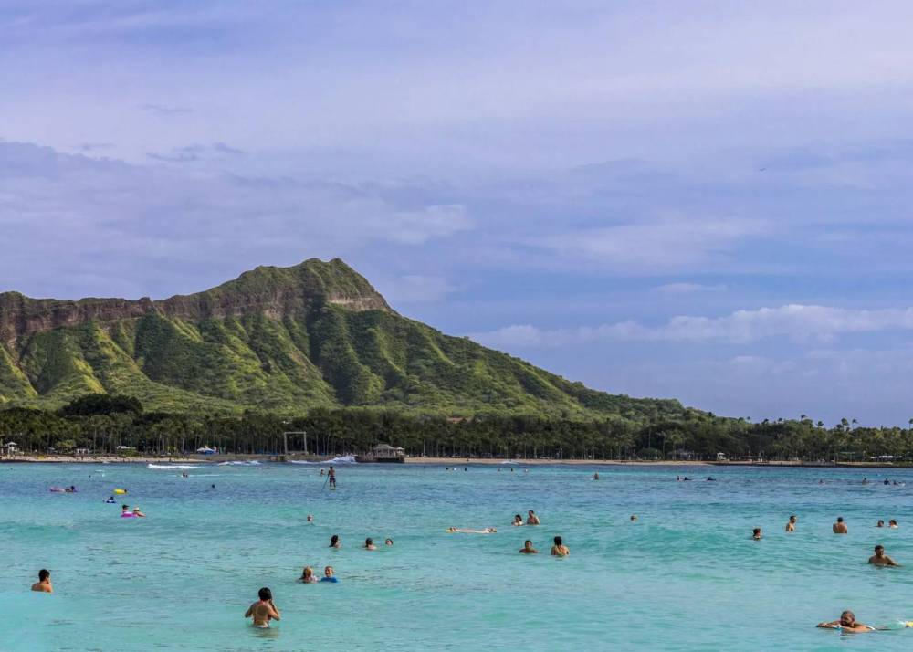 People swimming in clear ocean water with a green mountain in the background.