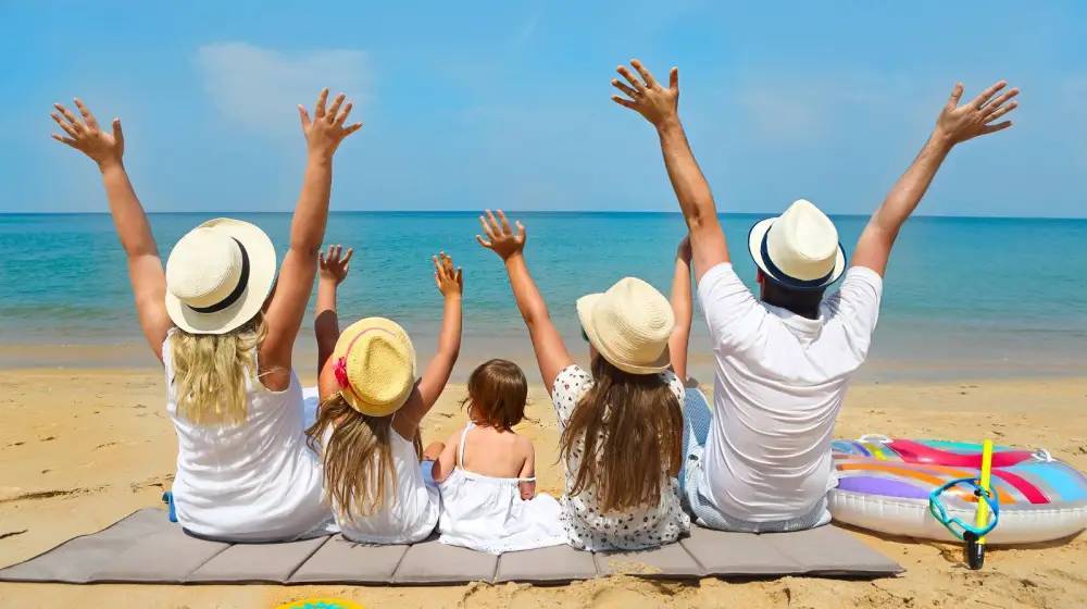 Family of five sitting on beach with arms raised, facing the sea.