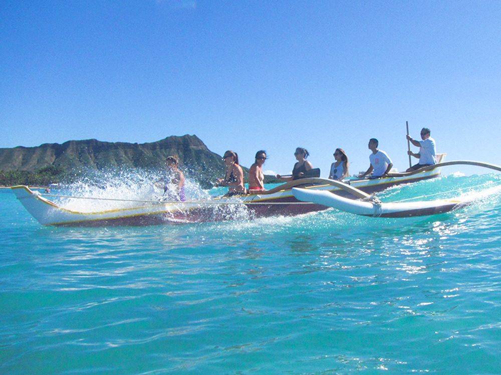 People paddling in outrigger canoe on blue ocean, mountain in background