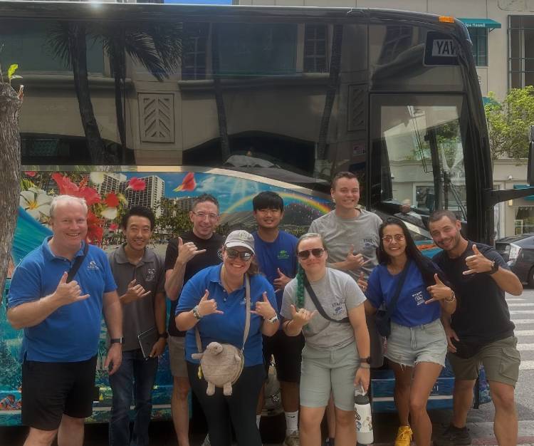 Group of eight people posing in front of a tour bus, making shaka signs.