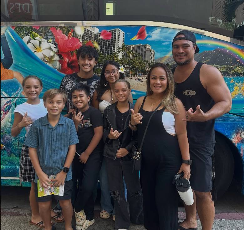 Group of smiling people posing in front of a colorful bus with Hawaiian-themed mural.