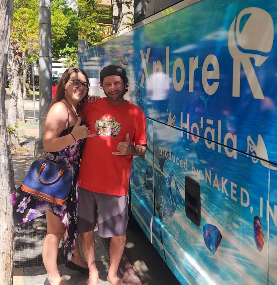 Two people smiling and gesturing thumbs up in front of a colorful Xplore tour bus.