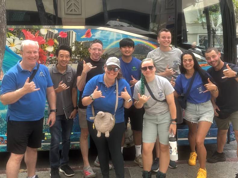 Group of nine people posing with hand signs in front of a colorful bus.