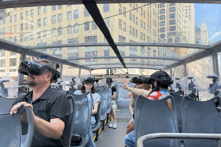 People on a glass-roofed bus wearing VR headsets in a city.