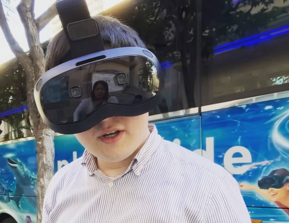 Person wearing VR headset outdoors, bus with blue graphics behind.