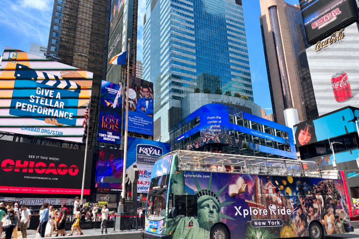 Double-decker tour bus in bustling city square with colorful billboards.