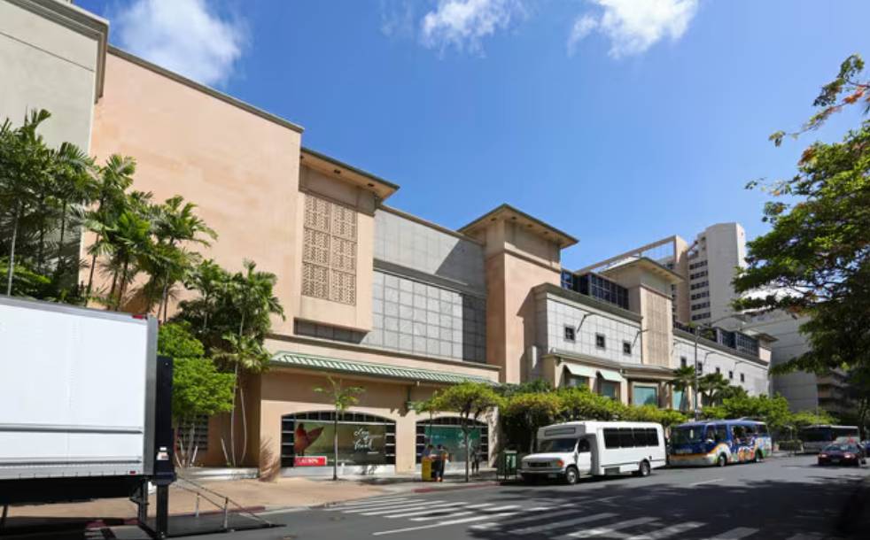 Street view of a large beige building with parked vehicles and trees.