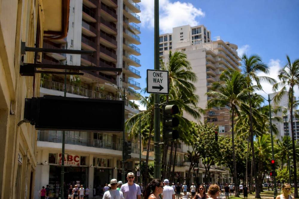 City street with people, palm trees, and tall buildings under a clear blue sky.